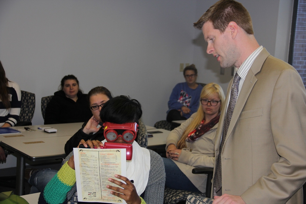A young woman in a college classroom using a vision simulator and goggles to read a menu